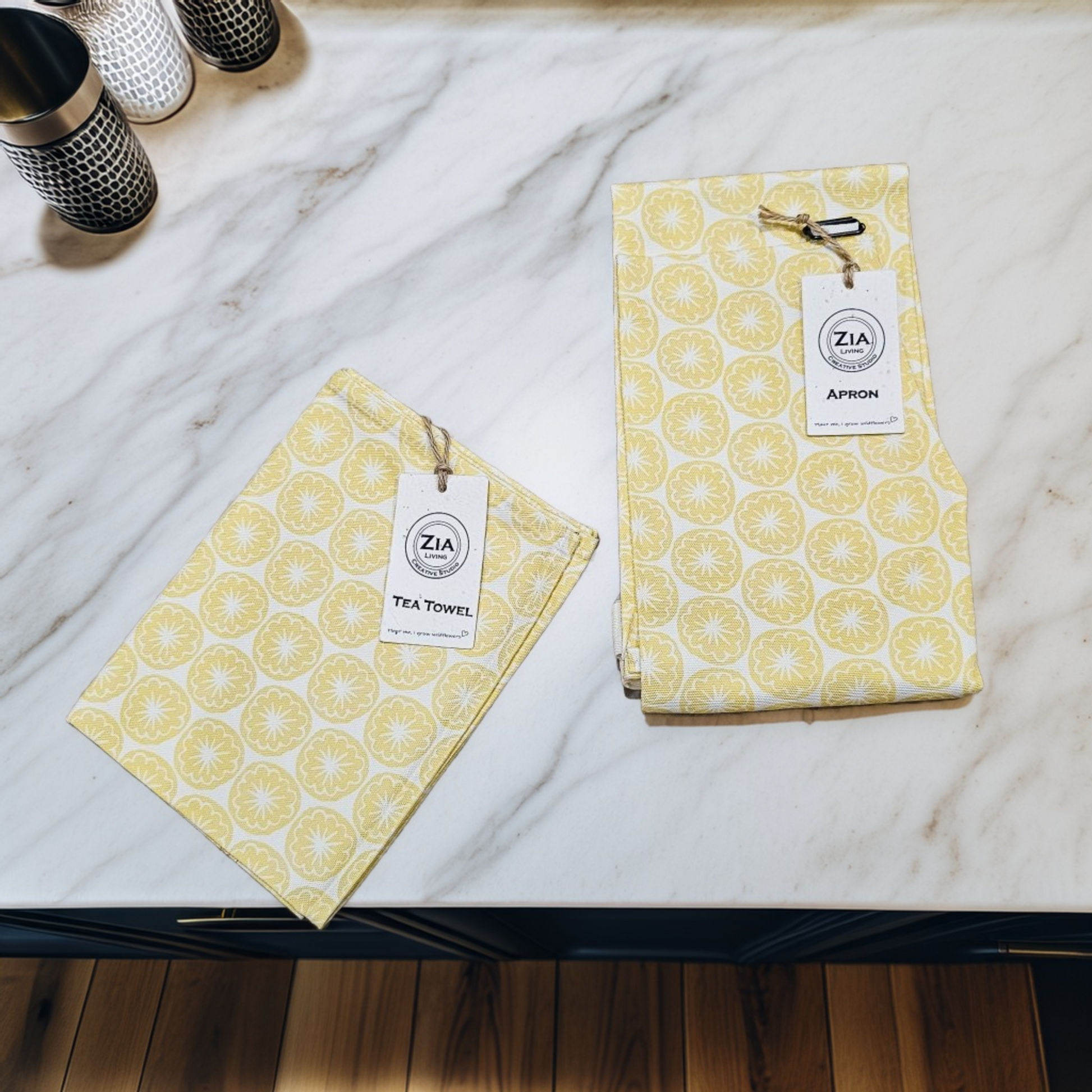  yellow Poppy Seed Head tea towel and apron from ZIA LIVING folded together on a marble surface with the wild flower seed card labels visible.