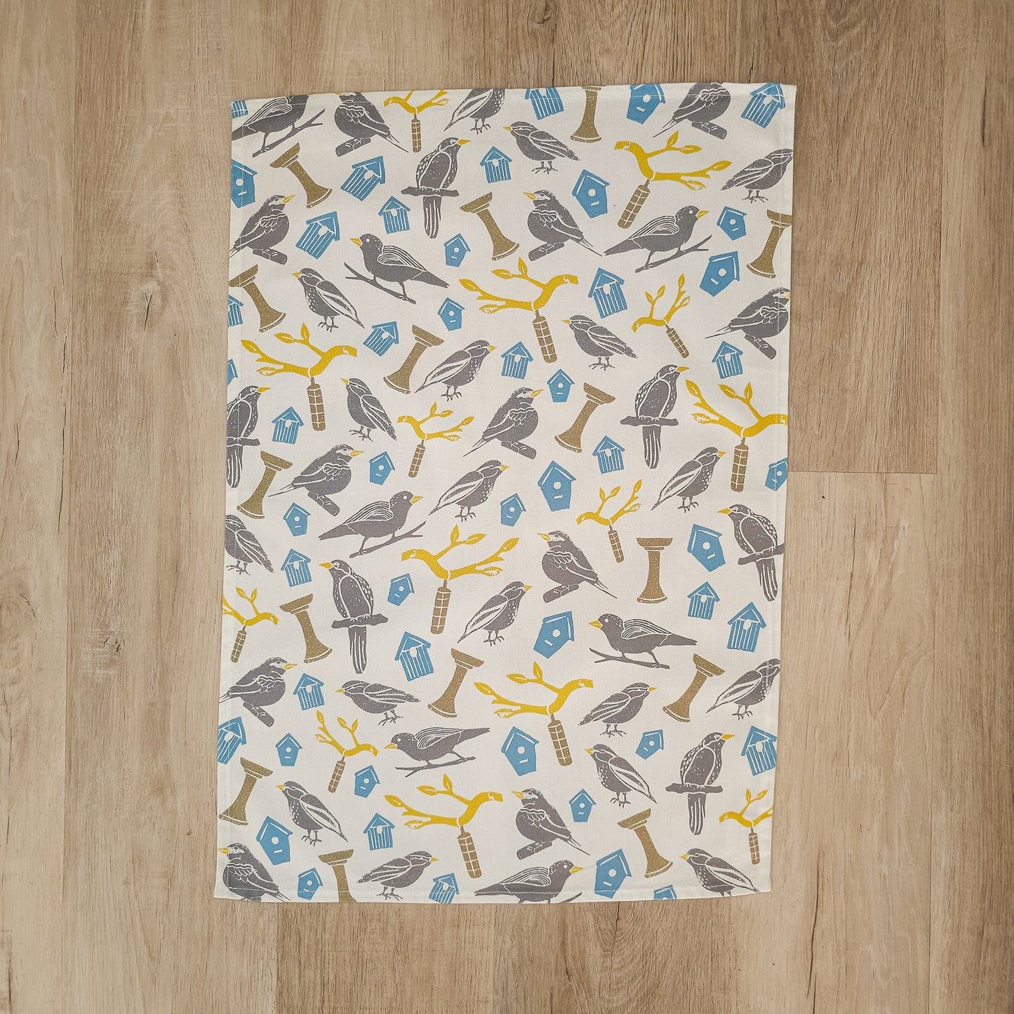 A Bird House print tea towel from ZIA LIVING laid on a wooden surface, showing the full bird and birdhouse pattern.