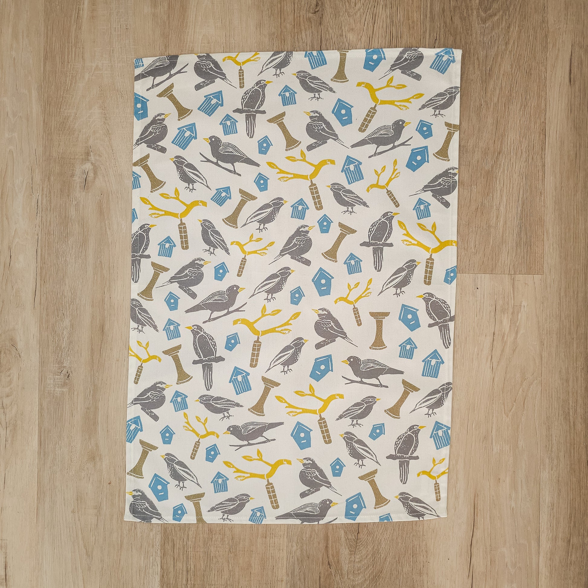 A Bird House print tea towel from ZIA LIVING laid on a wooden surface, showing the full bird and birdhouse pattern.