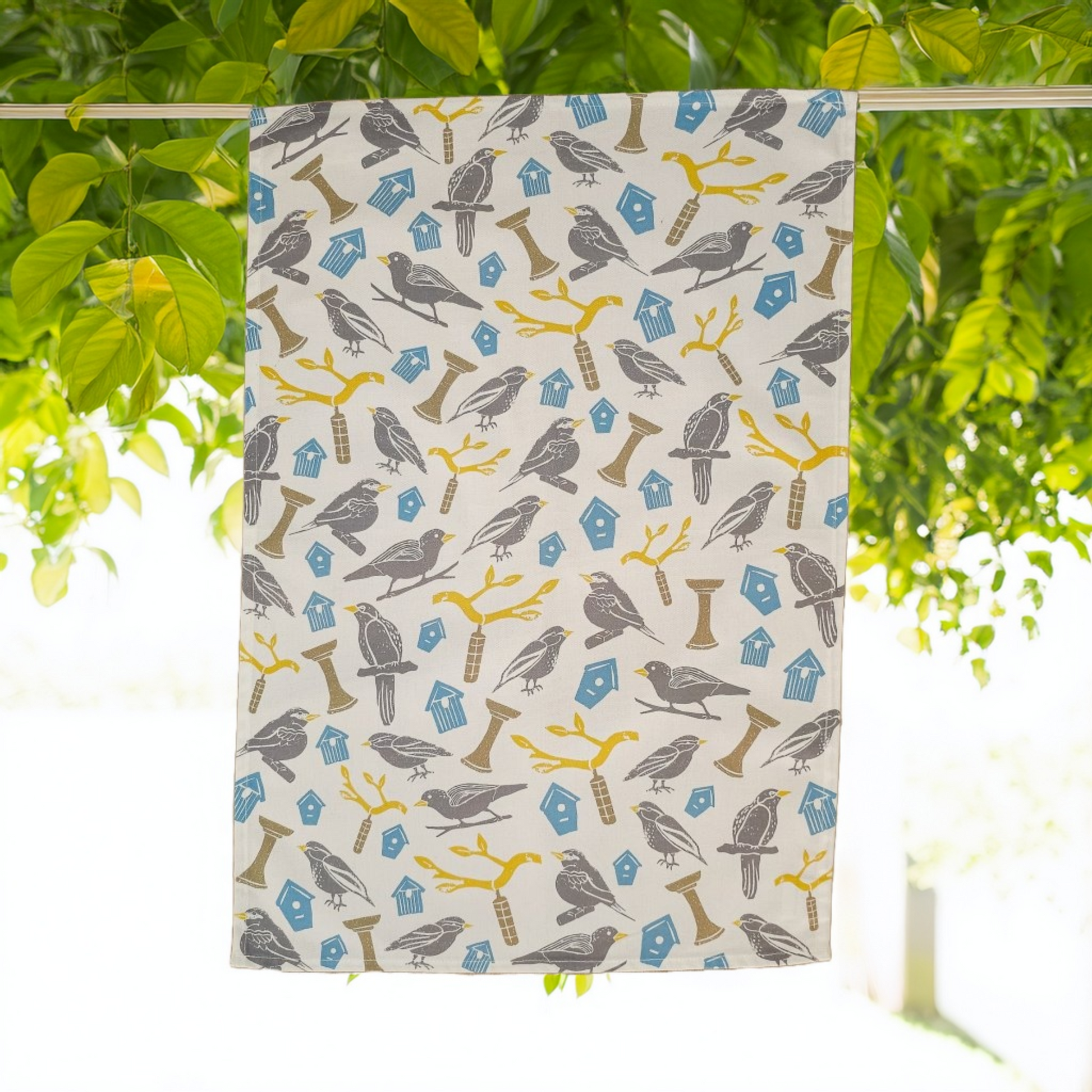 Patterned towel with birds and , yellow branches and bird houses hanging on a line with green leaves in the background designed by Zia Living