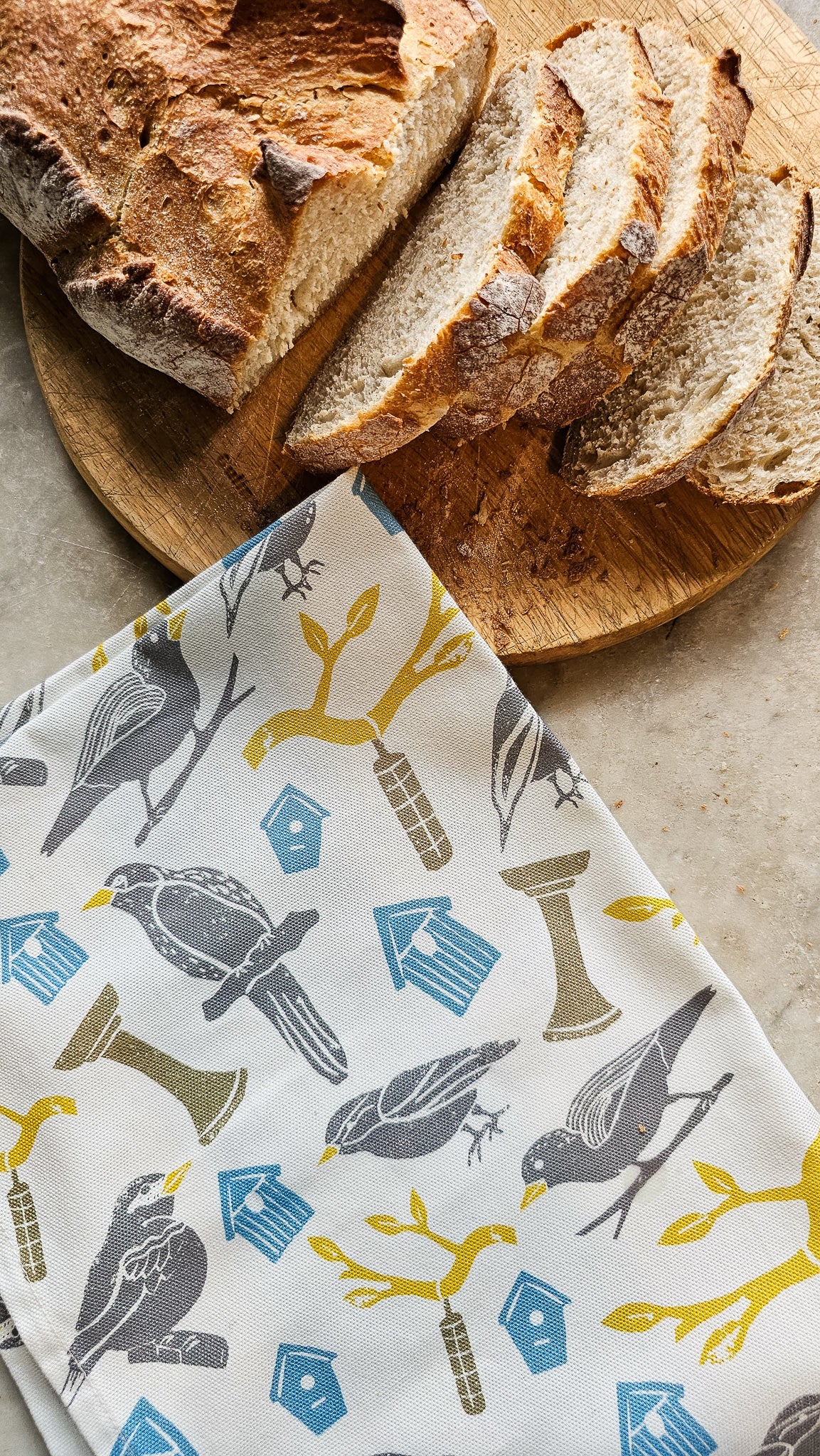 Loaf of bread on a wooden board with a Zia Living colourful bird print patterned cotton tea towel folded underneath