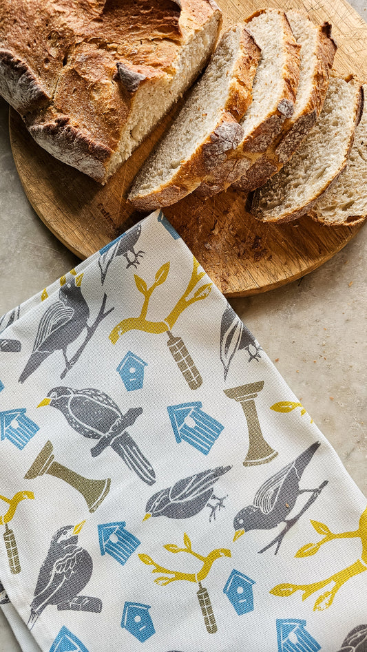 Loaf of bread on a wooden board with a Zia Living colourful bird print patterned cotton tea towel folded underneath