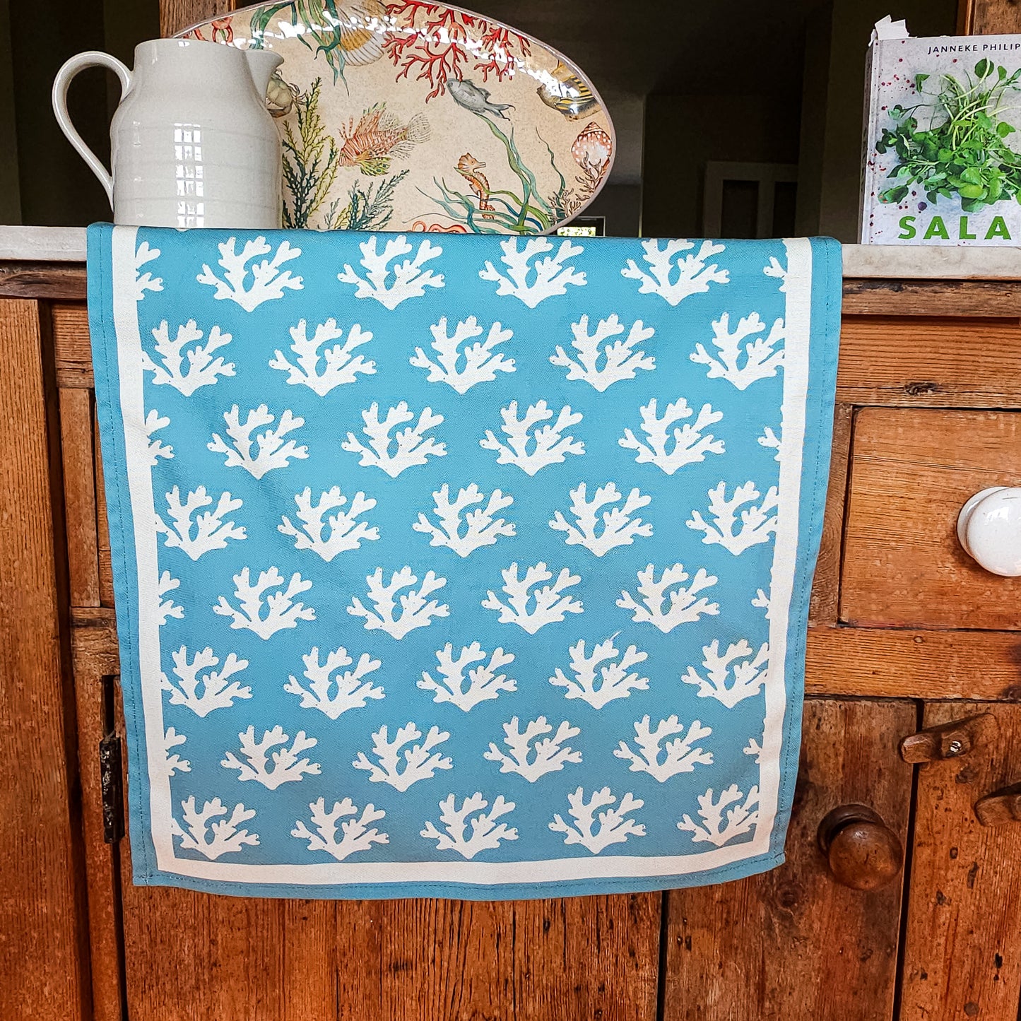 A blue Seaweed print tea towel from ZIA LIVING draped over a wooden cabinet door, with coastal‑themed ceramics and a cookbook displayed on top