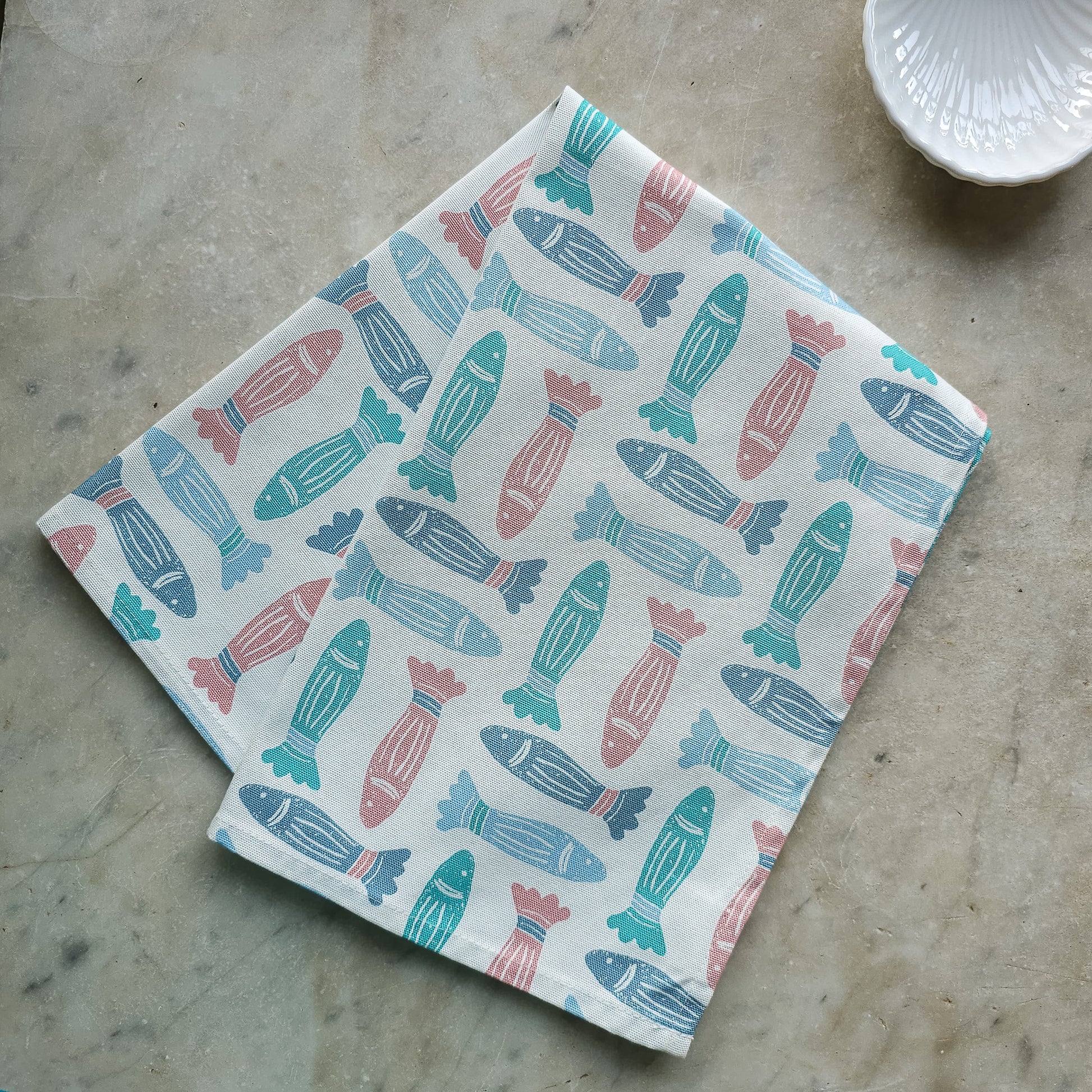 A blue, coral, and green fish‑print tea towel from ZIA LIVING folded on a textured  marble kitchen surface