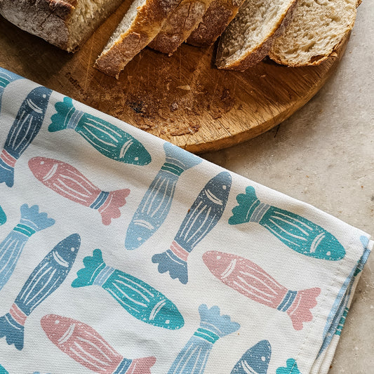 A blue, coral, and green fish‑print tea towel from ZIA LIVING folded next to sliced bread on chopping board