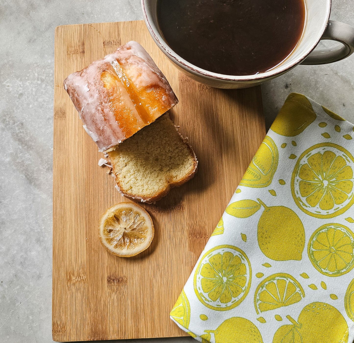 Lemon patterned tea towel folded beside a slice of iced lemon loaf and a cup of coffee on a wooden board — ZIA LIVING