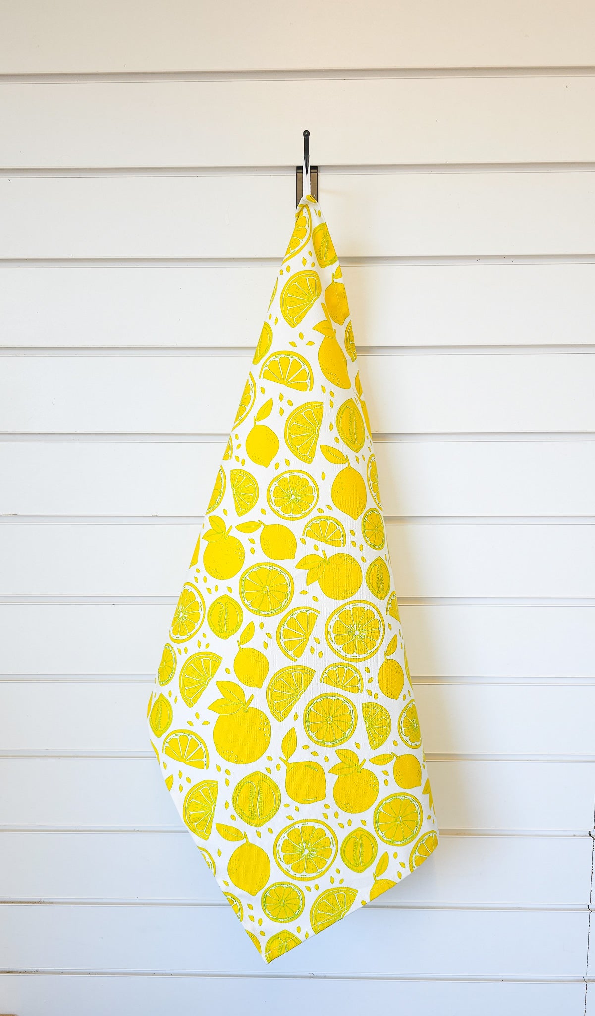 Lemon‑print tea towel hanging on a white wooden panel wall, featuring bright yellow lemons and slices in a fresh, summery design. ZIA LIVING