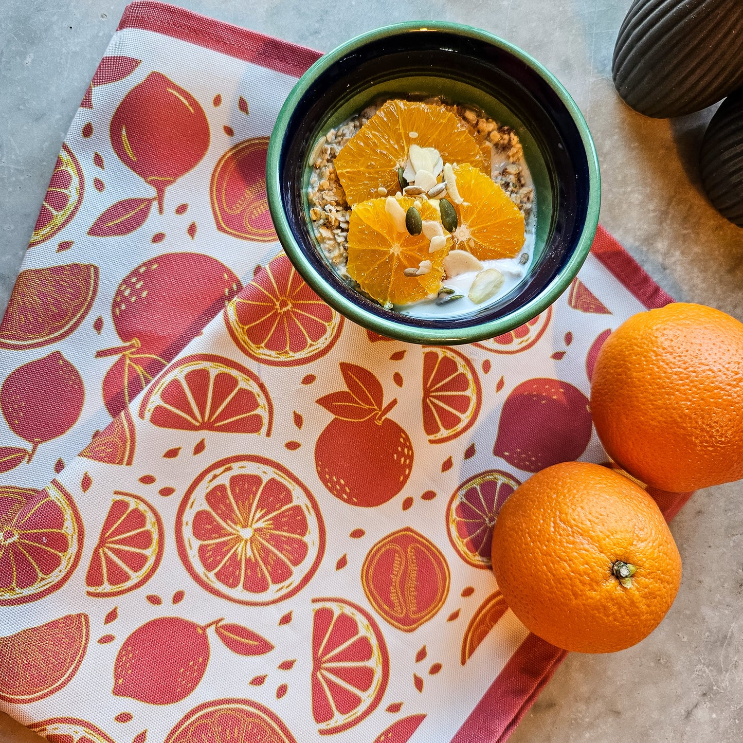 An orange and white orange‑print tea towel from ZIA LIVING laid beside a bowl of oatmeal topped with fresh orange slices on a textured surface.