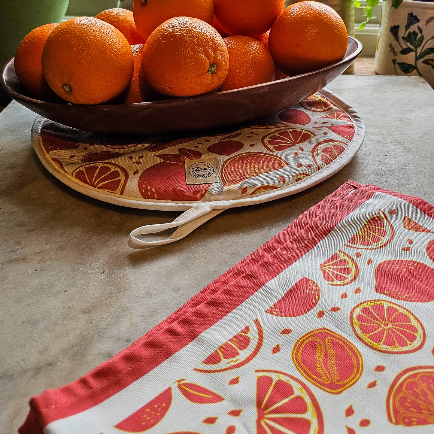 An orange‑and‑white citrus‑print tea towel from ZIA LIVING folded neatly beside a bowl of oranges on a decorative plate on marble worktop