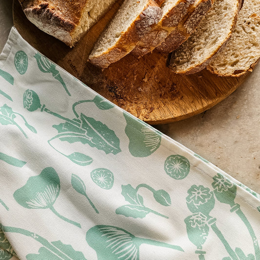 A mint‑green Wild Poppies tea towel from ZIA LIVING draped over a wooden board beside a sliced loaf of bread