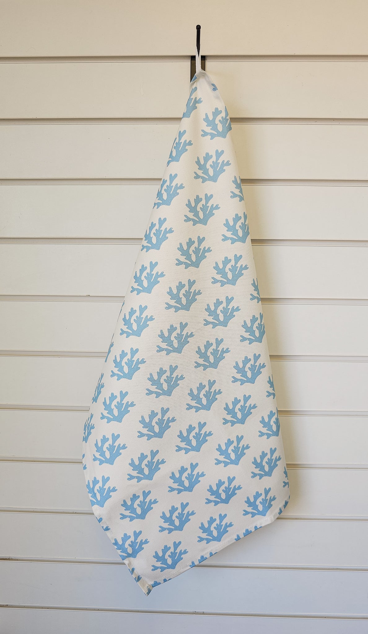 White tea towel with a soft blue seaweed coral pattern hanging on a black hook against a white panelled wall, styled in a calm coastal aesthetic. ZIA LIVING.