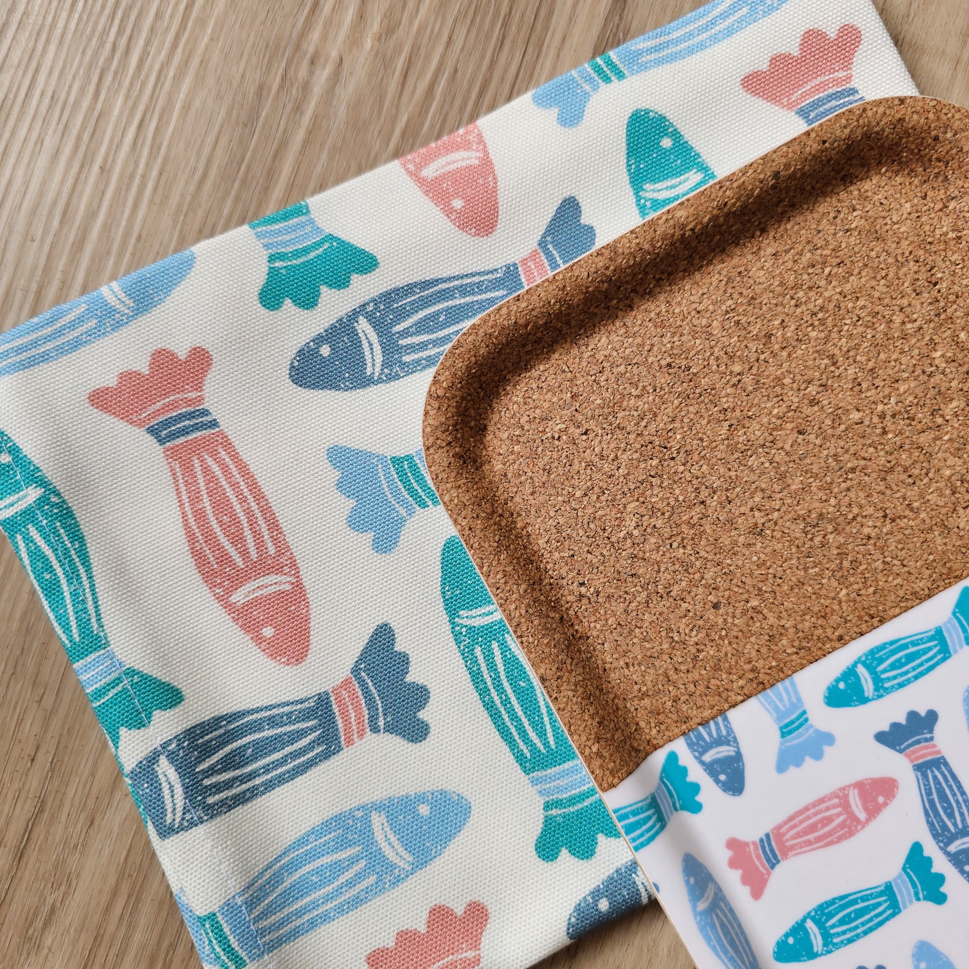 A blue, coral, and green fish‑print tea towel and matching tray with cork detail, from ZIA LIVING laid out on a wooden kitchen worktop