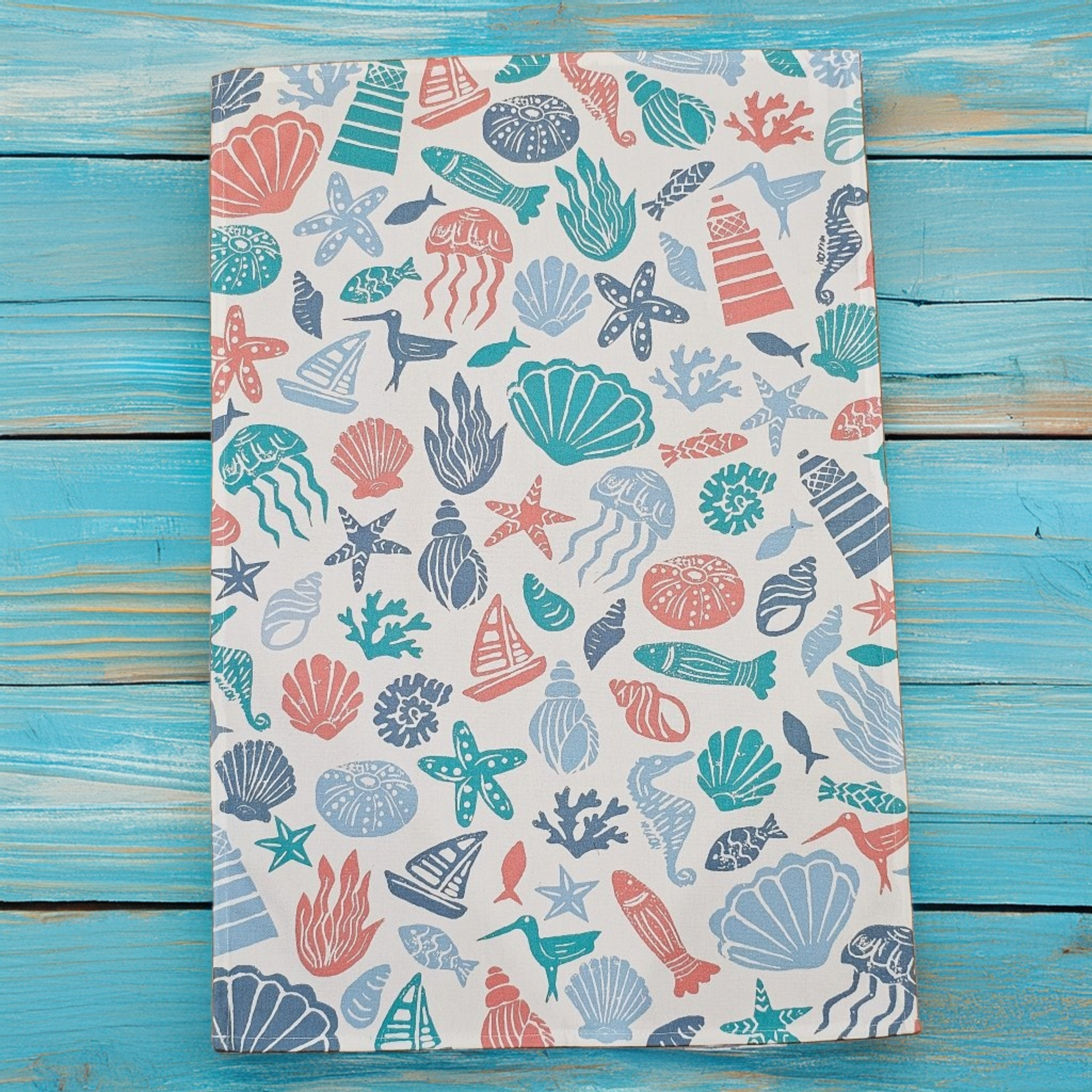 Under the Sea, a colourful coastal sea-themed tea towel folded on a vintage blue wooden surface in blue, coral and teal. Zia Living