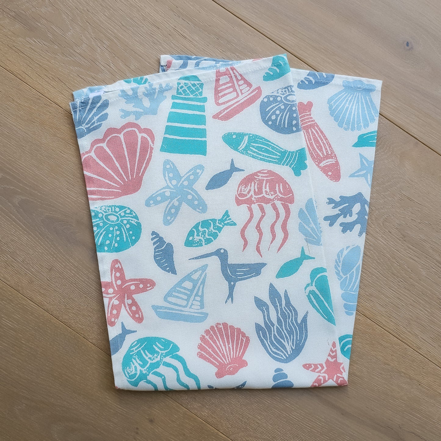 Under the Sea, a colourful coastal sea-themed tea towel folded on wooden surface in blue, coral and teal. Zia Living