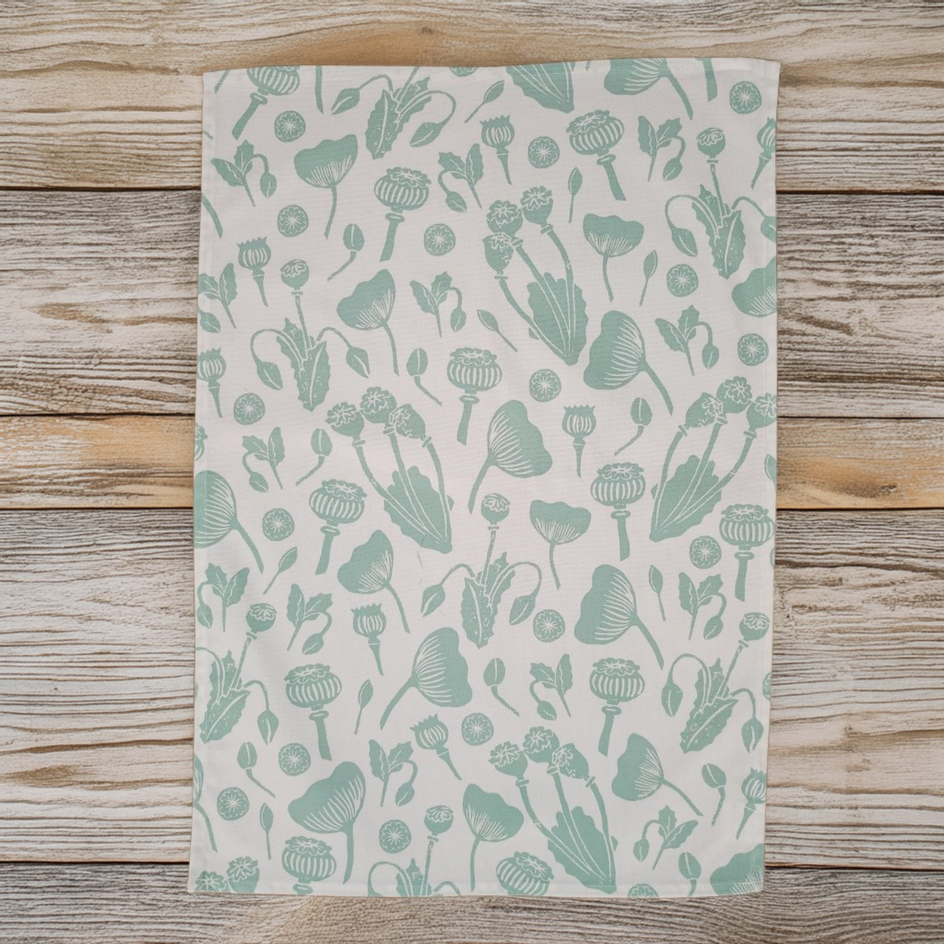 Wild Poppies botanical fabric laid flat on a wooden surface, featuring soft green floral and seed‑pod motifs on a light background — ZIA LIVING.
