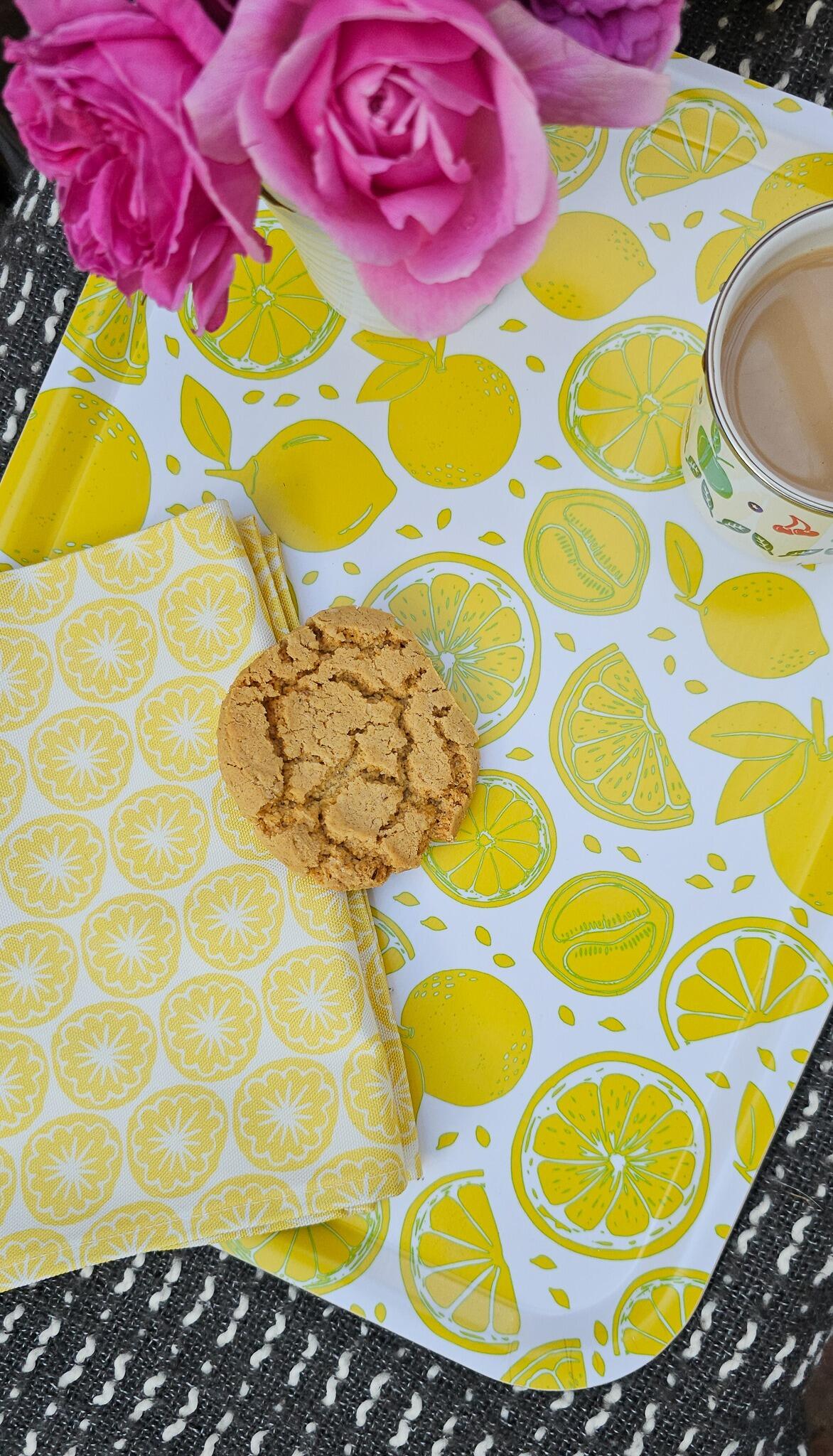 ZIA LIVING lemon‑print tray styled with a yellow seedhead tea towel, cookie, floral teacup, and pink roses on a woven brown‑and‑white surface.