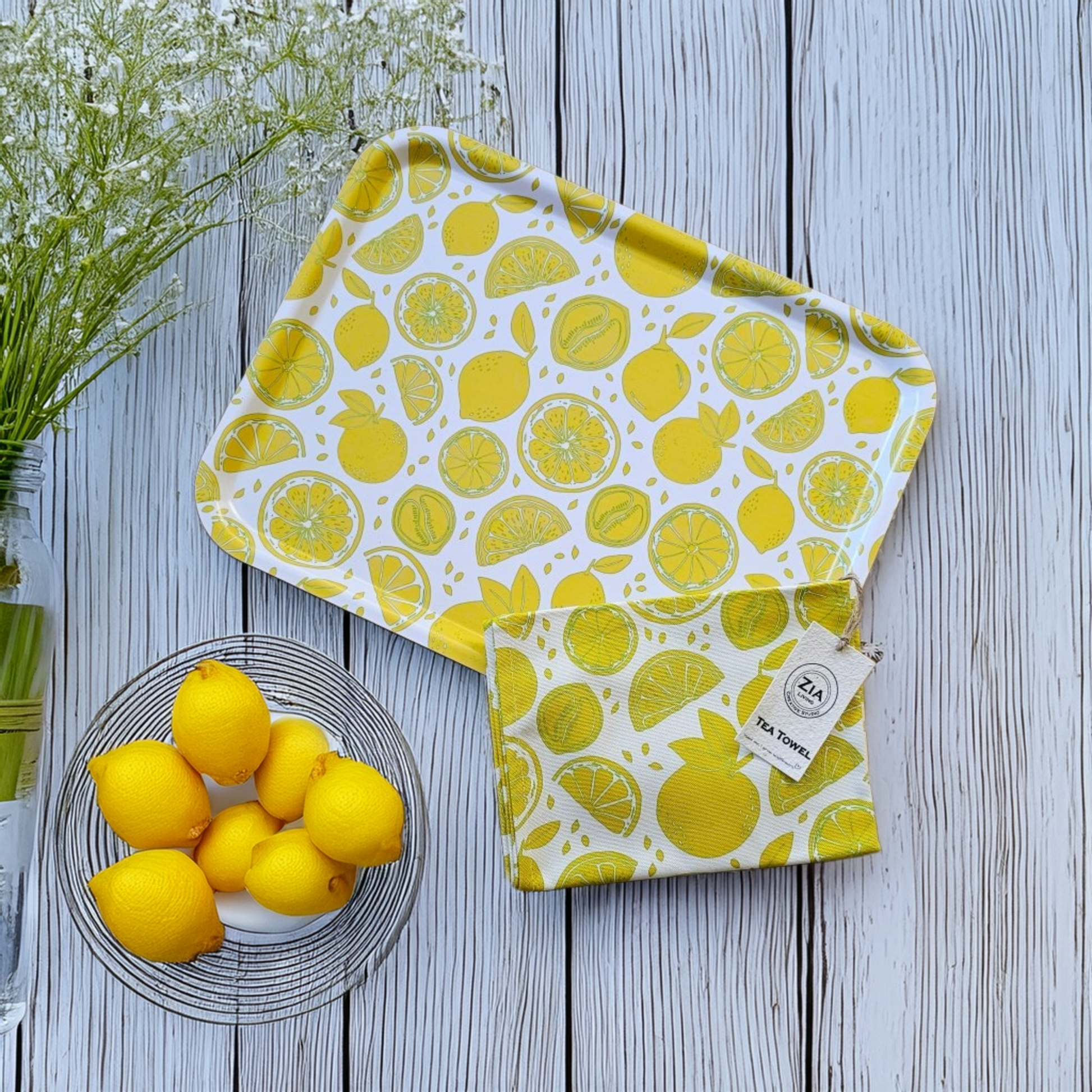 Lemon‑print rectangular tray styled with a matching lemon tea towel on a wooden surface, paired with a wire basket of fresh lemons and soft white florals for a bright, summery kitchen flat‑lay. ZIA LIVING citrus collection
