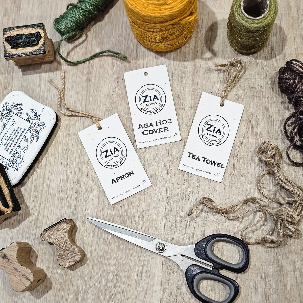 Three white paper tags from ZIA Handmade are laid out on a light wood surface with tea towel-making supplies around them, including yellow and green thread, scissors, wooden stamps, and twine. The tags read 'ZIA Handmade,' 'AGA Hob COVER,' 'APRON,' and 'TEA TOWEL.